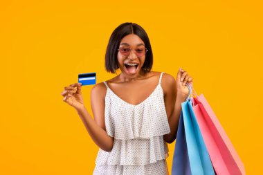 Yaz satışları. Alışveriş torbaları olan heyecanlı zenci kadın kredi kartını gösteriyor, turuncu arka planda OMG diye bağırıyor.