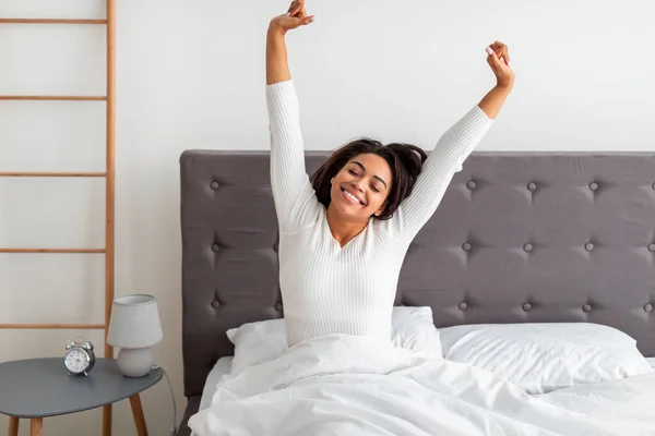Мирная черная женщина, вытягивающая руки и спину, сидит на кровати — стоковое фото