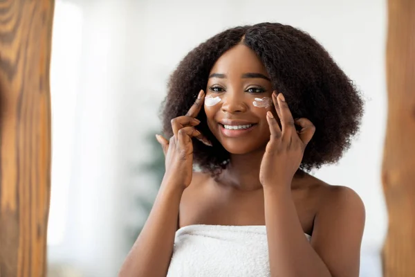 Atractiva mujer negra milenaria aplicando crema facial después de la ducha cerca del espejo en el interior. Concepto de spa y bienestar —  Fotos de Stock