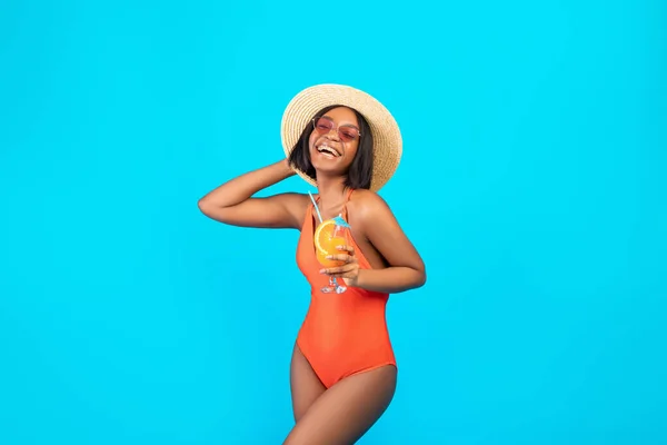 Fit dame noire en chapeau de paille et maillot de bain posant avec cocktail de fruits tropicaux pendant les vacances d'été, fond studio bleu — Photo