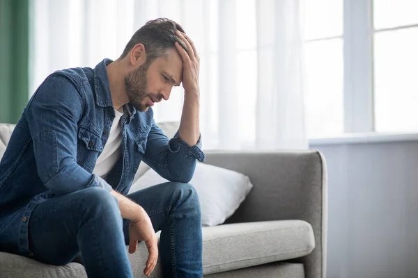 Uomo pensieroso seduto sul divano a casa, vista laterale — Foto Stock