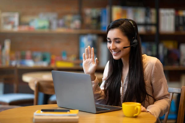 Emocionada señora árabe tutoría en línea en la cafetería, hablando y saludando a la cámara portátil a través de videoconferencia —  Fotos de Stock