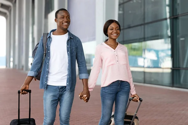 Retrato de alegre pareja negra sosteniendo las manos mientras camina cerca de la terminal del aeropuerto — Foto de Stock