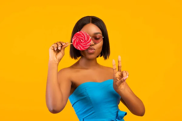 Femme noire en robe d'été et lunettes de soleil posant avec sucette, faire le geste V et les lèvres de sac sur fond orange — Photo