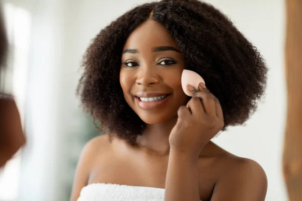 Mulher negra muito jovem com bela pele radiante aplicando fundação cosmética com aplicador perto do espelho dentro de casa — Fotografia de Stock