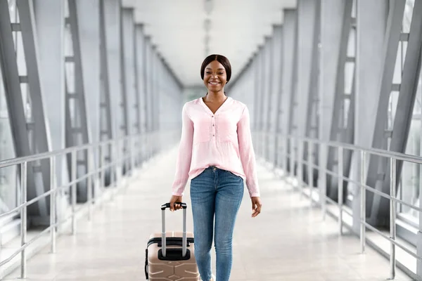 Letecká doprava. Krásná usmívající se mladá černoška kráčející na letišti se zavazadly — Stock fotografie