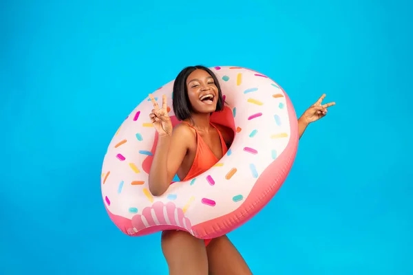 Mulher negra despreocupada em maiô vestindo anel de donut inflável, rindo e fazendo gesto de paz no fundo azul — Fotografia de Stock