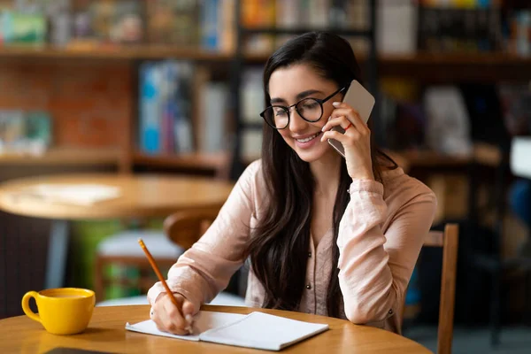 Joyeux arabe dame parler sur smartphone et prendre des notes à la cafétéria, gérer son horaire ou planifier des réunions. — Photo