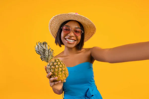 Sexy black lady in trendy summer dress taking selfie with pineapple on orange studio background — Stock Photo, Image