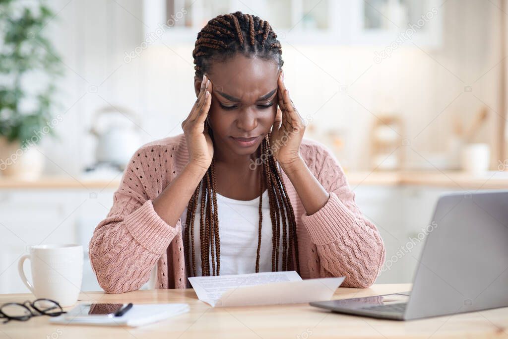 Stressed Black Woman Reading Documents While Sitting At Table In Kitchen