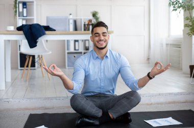 İş yeri stres yönetimi konsepti. Sakin bir Arap adam gözlerini kapatıp meditasyon yapıyor, modern ofiste yoga yapıyor.