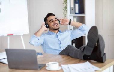 İşe ara verdim. Gülümseyen Arap adam kulaklıkla müzik dinliyor, iş yerinde dinleniyor.