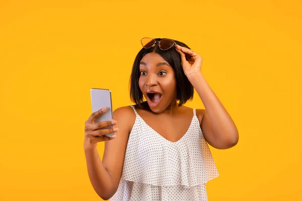 Потрясенная черная леди в летнем наряде с волнением смотрит на смартфон, поднимая солнечные очки на оранжевом фоне — стоковое фото