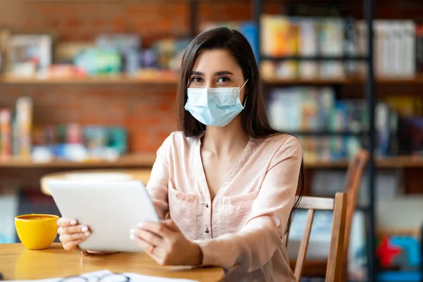 Latijnse vrouw met beschermend masker zittend in de coffeeshop en met behulp van digitale tablet, op zoek naar camera — Stockfoto