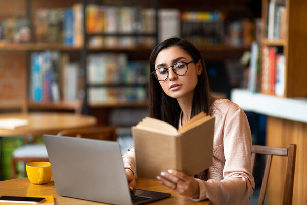 Задуманная латинская женщина читает книгу перед ноутбуком в кафетерии, учится и готовится к экзамену.