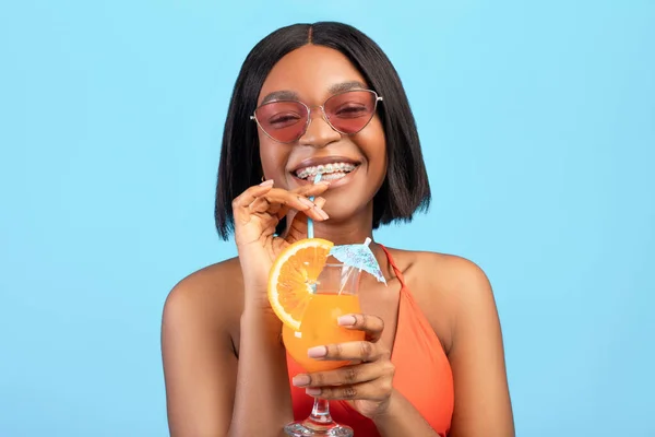 Portrait of cheerful black woman drinking refreshing cool fruit cocktail, having beach party on blue studio background — Stock Photo, Image