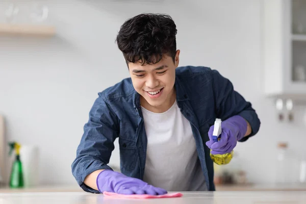 Веселий азіатський хлопець прибирає кухонний стіл з спреєм і тканиною — стокове фото