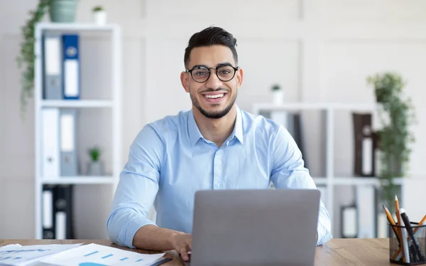 Glücklicher junger arabischer Geschäftsmann mit Brille sitzt am Schreibtisch und arbeitet im Büro am Laptop — Stockfoto