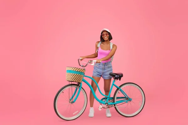 Positive young black lady in fashionable summer clothes posing with classic bicycle on pink studio background — Stock Photo, Image