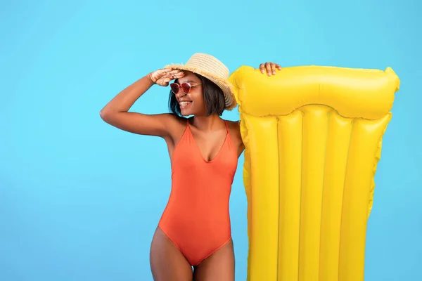 Joven dama negra en traje de baño con lilo inflable sosteniendo la mano por encima de sus ojos, mirando a la distancia sobre fondo azul — Foto de Stock