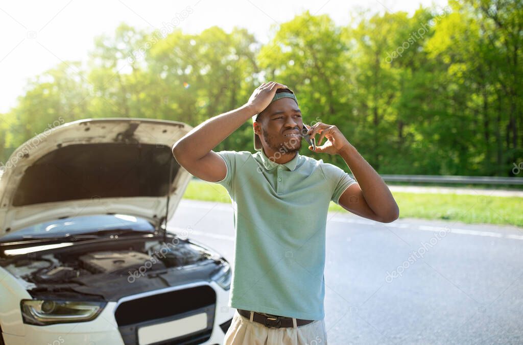 Unhappy black man standing near his broken car with open hood, calling breakdown road service on smartphone