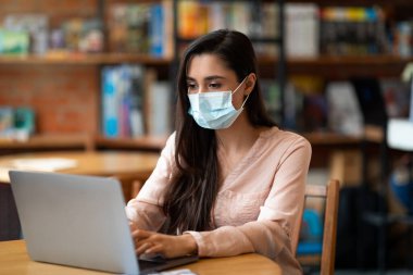 Yeni normal ve uzak çalışma. Laptop bilgisayarında maske takan Arap kadın, kafeden uzakta çalışıyor.