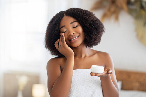 Beautiful young black woman applying face cream from jar, wearing bath towel, enjoying spa procedure at home