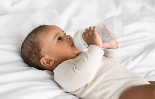 Χαριτωμένο μαύρο μωρό πίνοντας από το μπουκάλι που βρίσκεται στο κρεβάτι — Φωτογραφία Αρχείου