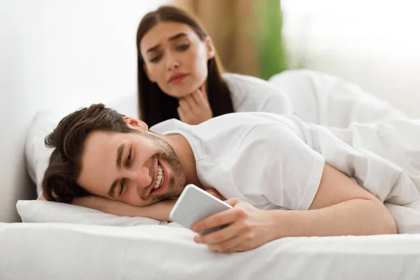 Esposa espiando mientras engaña marido mensajes de texto en el teléfono celular en el dormitorio — Foto de Stock