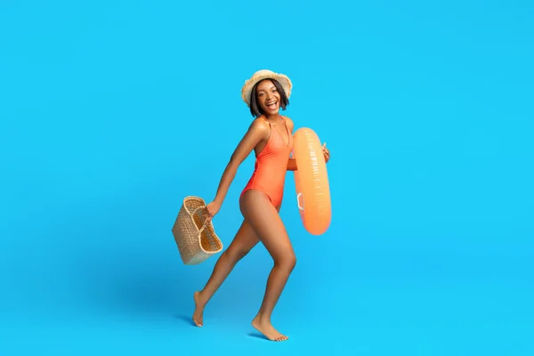 Plavání je zábava. Černá žena v plavkách jít do bazénu s nafukovací kroužek a sláma taška, modré pozadí, kopírovací prostor — Stock fotografie