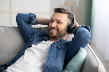 Kablosuz kulaklıkla kanepeye yaslanan mutlu sakallı adam.