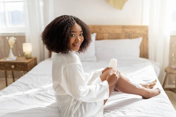 Bastante dama negra aplicando espuma de afeitar en sus piernas antes del procedimiento de depilación doméstica, mimándose en casa —  Fotos de Stock