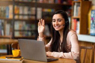 Kafede dinlenirken, bilgisayar ve kulaklık kullanarak video görüşmesi yapan heyecanlı Latin bayan, boşluğu kopyala