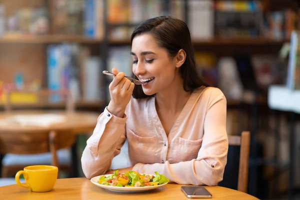Mujer latina feliz almorzando en la cafetería, disfrutando de deliciosa ensalada con los ojos cerrados y bebiendo bebida caliente. — Foto de Stock
