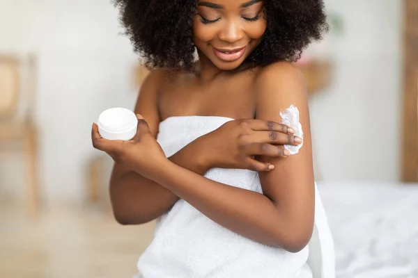 Beautiful black lady wearing towel after bath, applying cream, rubbing in nourishing body butter at home