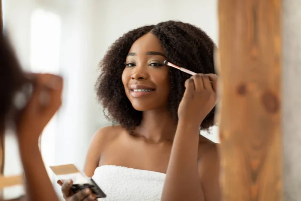 Wanita kulit hitam muda yang sensitif memakai eye shadow dengan pelamar di dekat cermin di rumah — Stok Foto