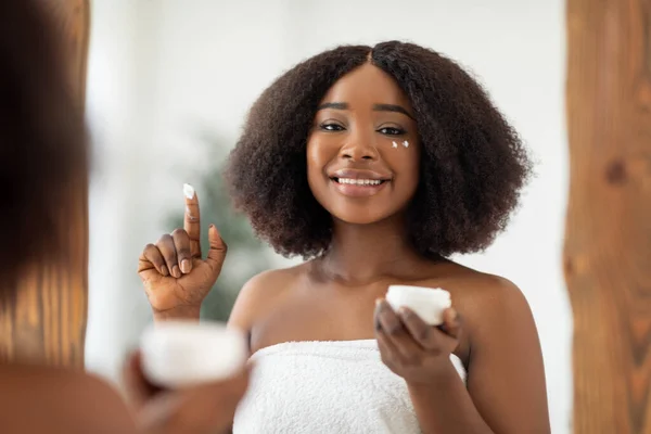 Biologische cosmetica voor een gezonde huid. Millennial zwarte dame aanbrengen gezichtscrème uit pot in de voorkant van spiegel thuis — Stockfoto