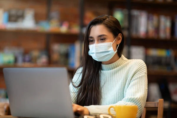 Удаленная работа во время пандемии ковида-19. Латиноамериканка в защитной маске печатает на ноутбуке в кафе — стоковое фото