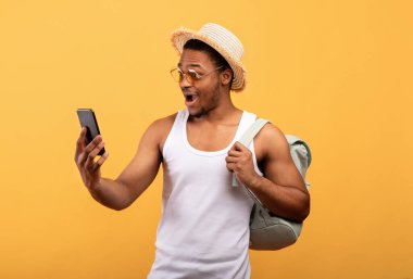 Sırt çantalı siyah bir adam heyecanlı bir şekilde akıllı telefona bakıyor. İnternetten son dakika turunu ayarlıyor. Sarı arka plan.