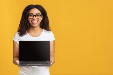 Mutlu Afrikalı Amerikalı kadın, sarı stüdyo arka planında boş bir dizüstü bilgisayarı kameraya gösteriyor.