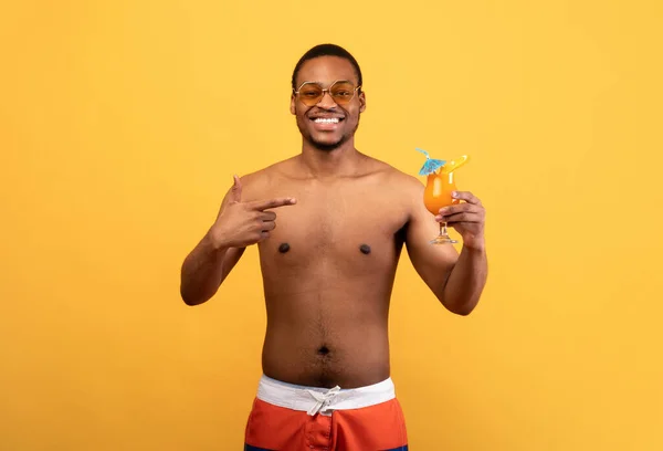 夏天很冷。身穿泳衣的快乐黑人指着水果鸡尾酒，对着黄色背景的相机微笑 — 图库照片
