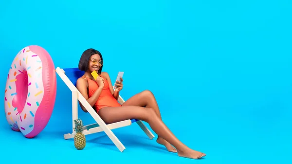 Mulher negra despreocupada segurando cartão de crédito e smartphone, compras on-line na cadeira lounge em fundo azul — Fotografia de Stock