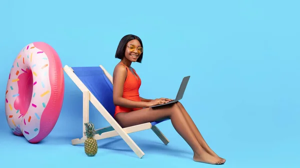 Trabalhando online no paraíso tropical. Mulher preta de biquíni sentada na cadeira lounge, usando laptop no fundo azul — Fotografia de Stock