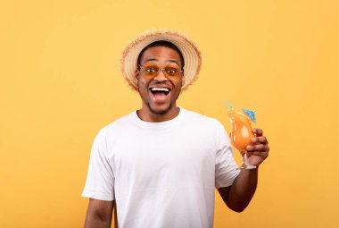 Neşeli siyah adam gündelik giysiler içinde ferahlatıcı bir yaz kokteyli tutuyor, sarı arka planda heyecan içinde ağzını açıyor.