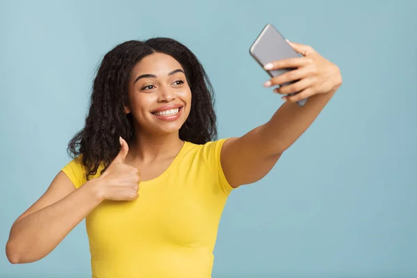 A mí me gusta. Feliz joven negro señora tomando selfie en el teléfono inteligente y mostrando el gesto de pulgar hacia arriba signo sobre fondo azul — Foto de Stock