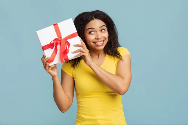 Překvapivý dárek. Nadšení černošky žena drží dárek, třesoucí se whapped box přes modré studio pozadí. — Stock fotografie