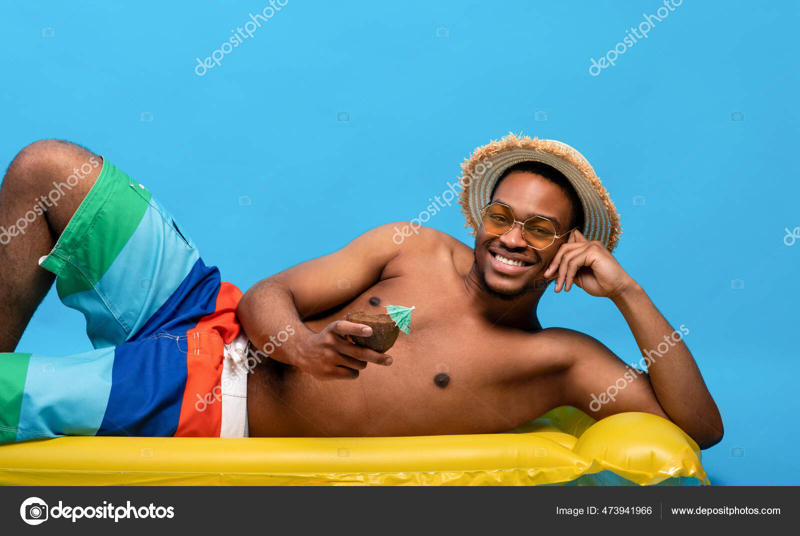 Funky černošky chlap v plavkách ležící na nafukovací lilo, pití letní  kokosový koktejl přes modré studio pozadí — Stock Fotografie © Milkos  #473941966