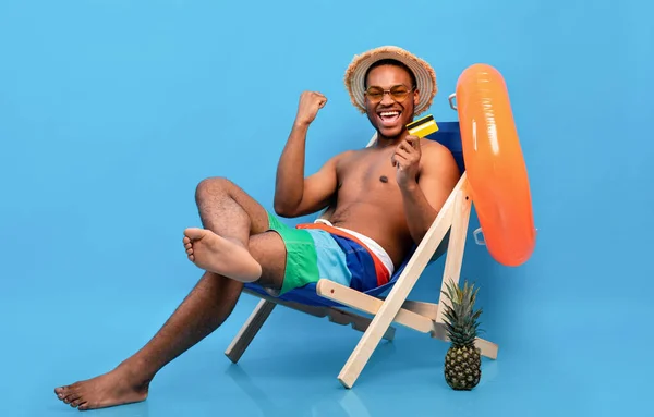 Presupuesto vacaciones de verano. Chico afroamericano alegre con tarjeta de crédito sentado en el sillón, haciendo un gesto SÍ —  Fotos de Stock