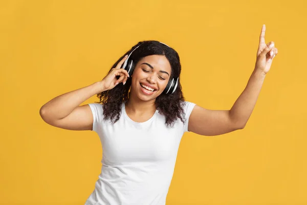 Amoureux de musique. Femme noire insouciante écoutant de la musique dans un casque et dansant sur fond de studio jaune — Photo