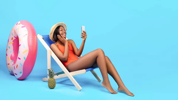 Mulher negra encantadora sentada na cadeira lounge, conversando com o amigo on-line no smartphone sobre fundo azul, espaço de cópia — Fotografia de Stock